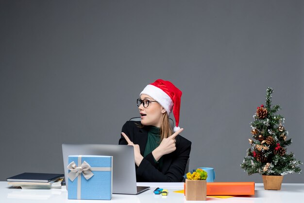 Positive Geschäftsfrau mit Weihnachtsmannhut, der an einem Tisch mit einem Weihnachtsbaum und einem Geschenk auf ihm auf dunklem Hintergrund sitzt