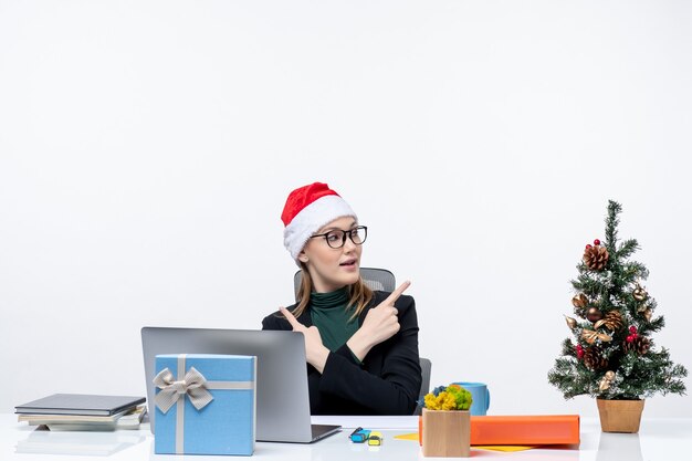 Positive Geschäftsfrau mit Weihnachtsmannhut, der an einem Tisch mit einem Weihnachtsbaum und einem Geschenk auf ihm auf weißem Hintergrund sitzt