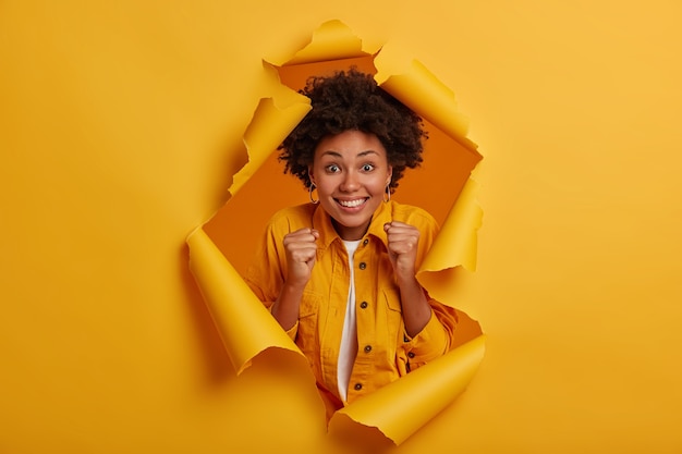 Positive glückliche Frau mit Afro-Frisur, hebt geballte Fäuste, lächelt breit, posiert im Hintergrund eines zerrissenen Lochs