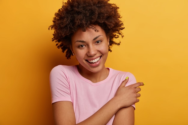 Positive gut aussehende lockige Frau mit lockigem Haar, lächelt angenehm, drückt gute Gefühle aus, hat einen charmanten Look, trägt ein lässiges Outfit und posiert über einer gelben Wand. Glückskonzept