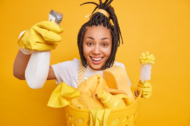 Positive Haushälterin trägt Schutzhandschuhe aus Gummi, die Hausreinigungsroutine hält Bürste und Waschmittel sammelt schmutzige Wäsche einzeln auf gelbem Hintergrund. Zimmerreinigung am Reinigungstag