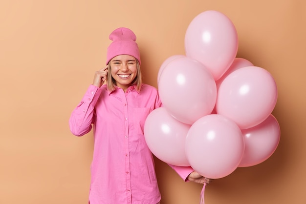 Positive, hellhaarige junge frau vermeidet laute musik auf partystöpseln, lächelt und zwinkert augen in rosafarbener kleidung posiert mit aufgeblasenen luftballons vor braunem hintergrund. hier ist es zu laut.