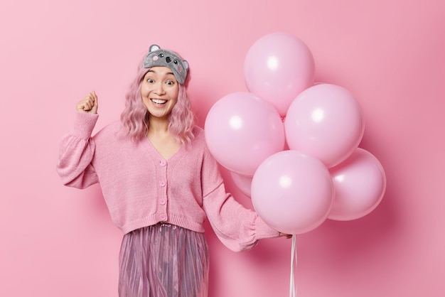 Positive hübsche asiatische Frau mit rosigen Haaren wartet auf besondere Anlässe, lächelt gerne und trägt Schlafmaskenpullover und Faltenrock hält einen Haufen aufgeblasener Luftballons isoliert über rosa Hintergrund