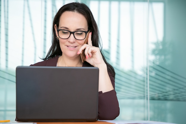 Positive hübsche Geschäftsfrau in den Gläsern, die auf Handy sprechen, am Computer im Büro arbeiten, Laptop am Tisch verwenden