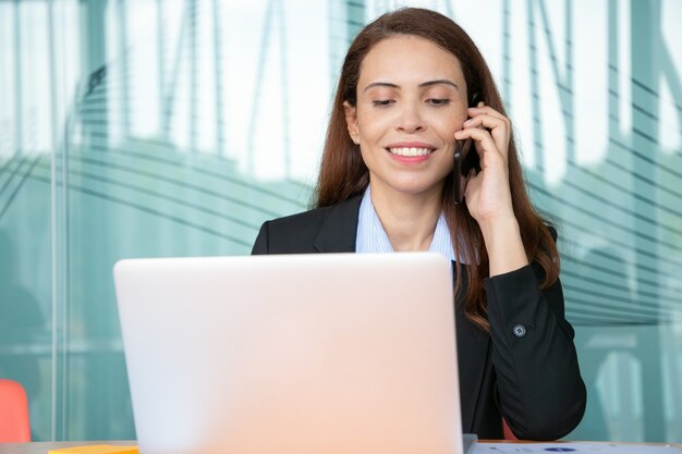 Positive hübsche junge Geschäftsfrau, die auf Handy spricht und lächelt, am Computer im Büro arbeitet, Laptop am Tisch benutzt