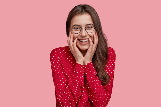 Positive junge Dame mit sanftem Lächeln, trägt eine Brille, modisches Hemd, zeigt natürliche Schönheit