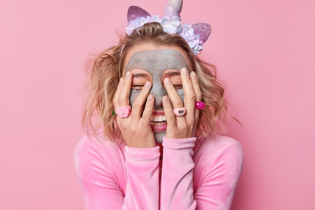 Kostenloses Foto positive junge europäerin bedeckt gesicht mit händen kichert positiv trägt einhorn-stirnband und pullover trägt schönheits-ton-maske zur hautbehandlung isoliert über rosa hintergrund auf wellness-konzept