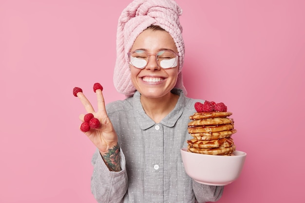 Positive junge Frau hat Spaß beim Essen leckerer Pfannkuchen mit Himbeeren und Honig macht Friedensgeste lächelt froh trägt Pyjama und Handtuch auf dem Kopf Posen vor rosa Hintergrund bleibt glücklich