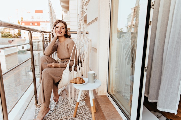Positive junge Frau im langen Kleid, das am Balkon mit Kaffee und Croissant sitzt. Foto des barfüßigen lockigen Mädchens, das Frühstück an der Terrasse genießt.