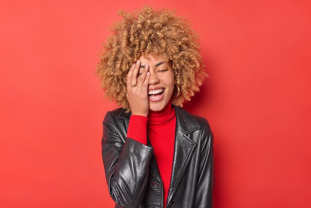 Kostenloses Foto positive junge frau mit blonden locken lässt die gesichtspalme überglücklich lachen, trägt rollkragenpullover und schwarze lederjacke einzeln auf leuchtend rotem hintergrund. aufrichtige authentische emotionen