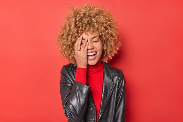 Positive junge Frau mit blonden Locken lässt die Gesichtspalme überglücklich lachen, trägt Rollkragenpullover und schwarze Lederjacke einzeln auf leuchtend rotem Hintergrund. Aufrichtige authentische Emotionen