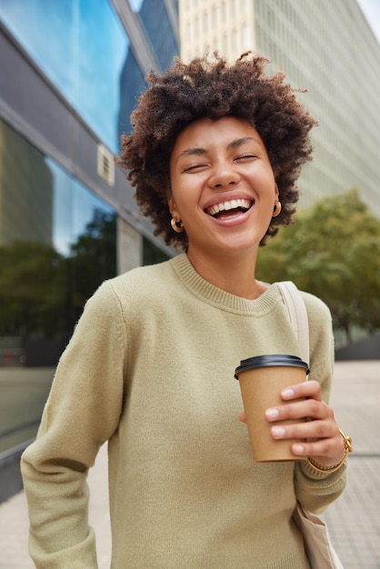 Positive junge Frau mit fröhlichem Ausdruck lächelt glücklich und hört etwas Lustiges Getränke zum Mitnehmen Kaffee, gekleidet in lässigen Pullover-Posen vor unscharfem Hintergrund. Menschen Lifestyle und Freizeitkonzept.
