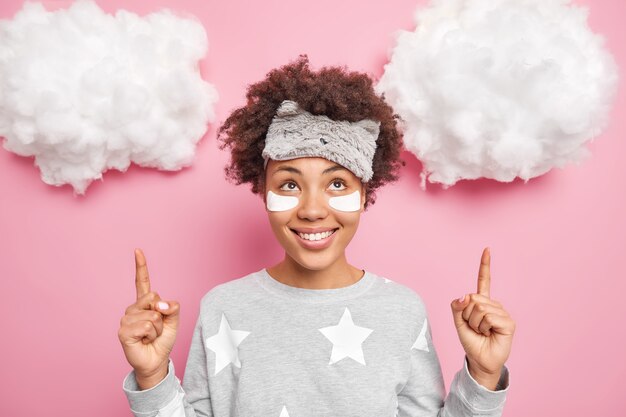Positive junge Frau mit lockigem Haar trägt Schlafmaske und Schlafanzug trägt Schönheitsflecken unter den Augenpunkten oben auf weißen Wolken auf. Das Produkt zum Schlafen hat gute Laune