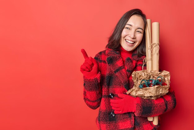 Positive junge Frau trug Papierrollen zum Verpacken von Geschenken und Retro-Girlande zum Dekorieren des Neujahrsbaums in Wintermantelhandschuhen zeigt auf leeren Kopierraum vor rotem Hintergrund