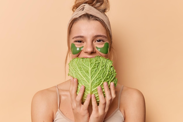 Positive junge Frau verwendet Naturprodukte für die Hautpflege hält Kohl über dem Mund trägt grüne Hydrogel-Patches unter den Augen trägt Stirnband und T-Shirt einzeln auf beigem Hintergrund. Schönheitszeit