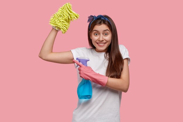 Positive junge Frau zeigt Muskeln nach müder Arbeit über Haus, gekleidet in weißes lässiges T-Shirt, hält Flasche Spray