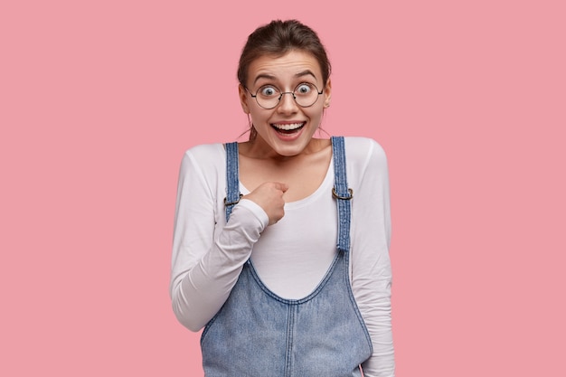 Positive kaukasische Frau zeigt auf sich selbst, sieht freudig aus, fragt, ob sie eine echte Kandidatin für den Gewinn des Wettbewerbs ist, trägt Denim-Sarafan und eine runde Brille