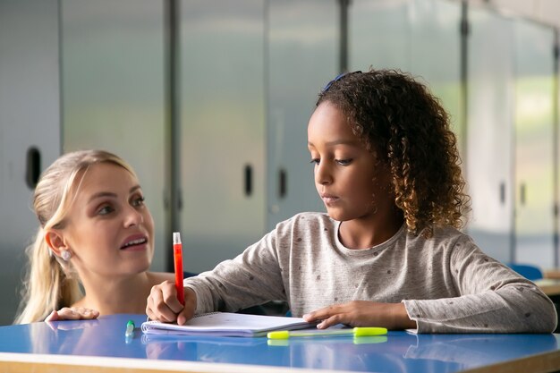 Positive Lehrerin, die lockigen Mädchen hilft, ihre Aufgabe zu erfüllen