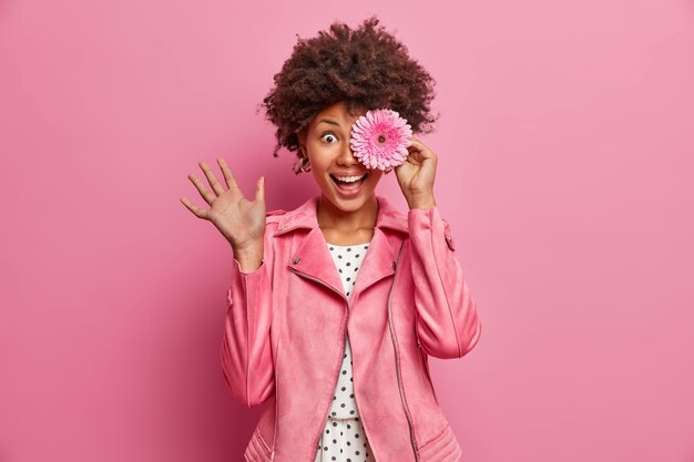 Positive lockige Frau hat Floristenkarriere, trägt rosa Gerbera, bedeckt Auge mit Blume, gekleidet in stilvolle Jacke, posiert drinnen, töricht herum, genießt angenehmen Geruch. Blüte, Duft