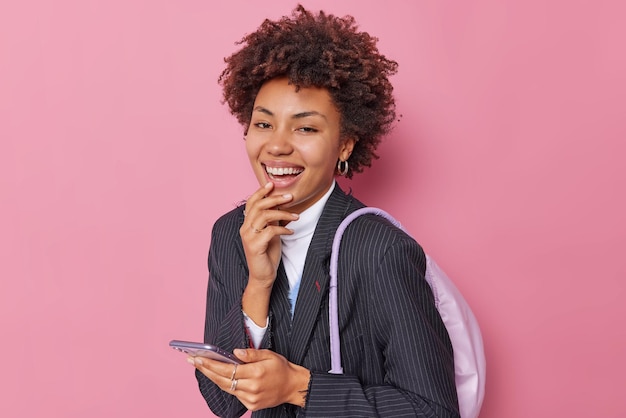 Positive lockige frau in eleganter schwarzer jacke trägt rucksack verwendet smartphone zum senden von textnachrichten lacht freudig isoliert über rosafarbenem hintergrund. glücklicher technologiebenutzer mit gadget