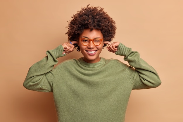 Kostenloses Foto positive lockige frau lächelt glücklich stecker ohren vermeidet sehr laute musik trägt transparente brille und pullover hat fröhliche stimmung über braune wand isoliert