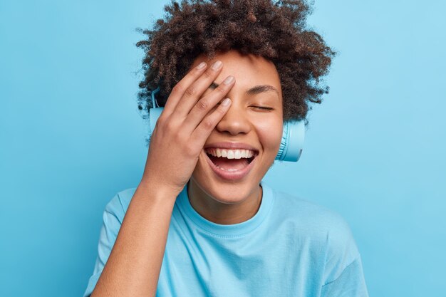 Positive lockige Frau lässt Gesicht Palme glücklich lächeln hat sorglosen Ausdruck hört Audiospur über Kopfhörer gekleidet in lässigem T-Shirt isoliert über blauer Wand. Emotionen Lebensstil