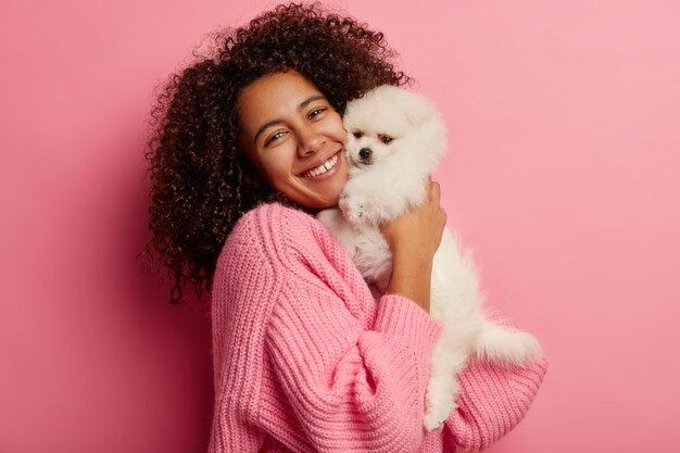 Positive lockige Frau umarmt kleinen Welpen, drückt zärtliche Gefühle zum Haustier aus, gekleidet in gestrickten Pullover, hatte Besuch beim Groomer, posiert vor rosa Hintergrund.