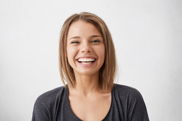 Kostenloses Foto positive menschliche gesichtsausdrücke und emotionen. fröhliches attraktives junges mädchen mit breit grinsender bob-frisur, die ihre perfekten weißen zähne zeigt, während sie schöne zeit drinnen verbringt