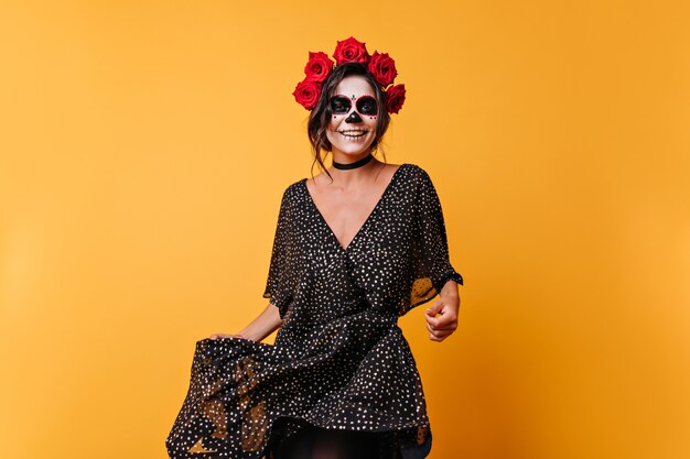 Positive mexikanische Frau, die mit Lächeln auf gemaltem Gesicht tanzt. Porträt des hübschen Mädchens mit gewelltem Haar im orangefarbenen Studio.