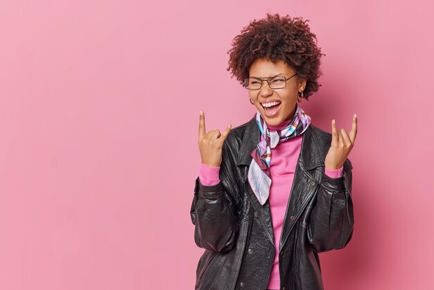 Positive, modische, lockige Frau macht Rock'n'Roll-Gesten, ruft laut aus, genießt Partyzeit, Lieblingsmusikshows, Heavy-Metal-Schild, trägt Lederjacke und Kopftuch, isoliert auf rosa Hintergrund