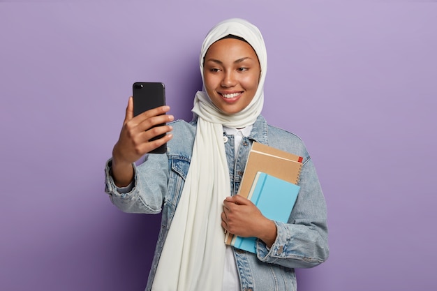 Positive muslimische Frau mit angenehmem Lächeln, nimmt Selfie auf modernem Smartphone, steht mit Spiralblock und Lehrbüchern isoliert auf lila Studiowand