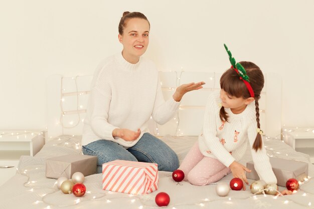 Positive Mutter und Tochter tragen weiße Pullover im Casual-Stil, die auf dem Bett sitzen und mit Weihnachtskugeln dekorieren, Frau mit erhobenem Arm und lächelnd in die Kamera.