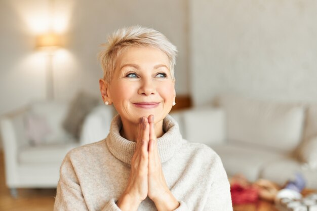 Positive reife europäische Frau in warmem Pullover mit verträumtem, erstauntem Gesichtsausdruck, der die Hände zusammenpresst und lächelt, auf das Beste hofft und Gott um Gesundheit und Wohlbefinden bittet. Glaubenskonzept