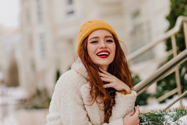 Positive rothaarige Frau, die über verschwommene Natur lacht. Raffiniertes Ingwermädchen, das während des Winterfotoshootings lächelt.