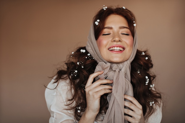 Positive rothaarige Frau lacht mit geschlossenen Augen. Porträt der Frau im beige Schal und mit den weißen Blumen im Haar.