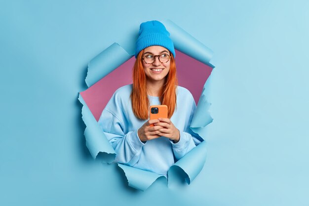 Positive rothaarige Teenager-Mädchen-Technologie Benutzer hält Handy sendet Textnachrichten Blicke beiseite trägt gerne blauen Hut und Pullover sieht nachdenklich beiseite bricht durch blaues Papierloch