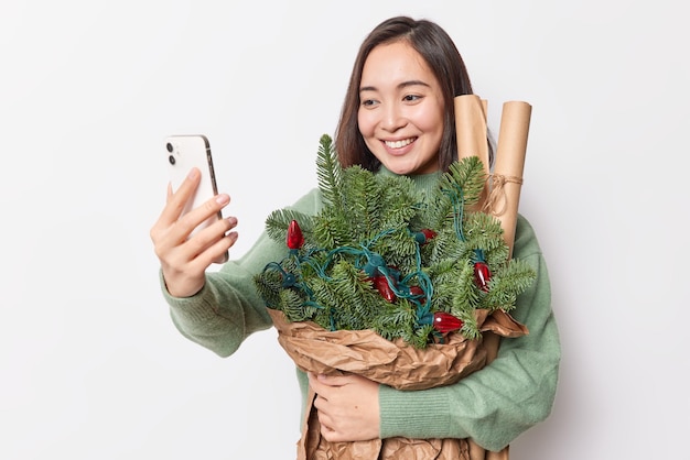 Positive schöne asiatische Frau macht Selfie auf dem Smartphone und hält Fichtenzweige in Papier gewickelt, das mit Retro-Girlanden geschmückt ist, lächelt glücklich isoliert über weißem Hintergrund. Weihnachtsvorbereitung