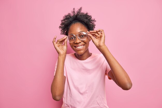 Positive schöne Frau mit lockigem Haar, dunkle Haut berührt den Rand der Brille, lächelt zahnig und blickt direkt in die Kamera, die fröhlich in einem lässigen T-Shirt gekleidet ist, das über rosafarbenem Hintergrund isoliert ist Emotions-Konzept