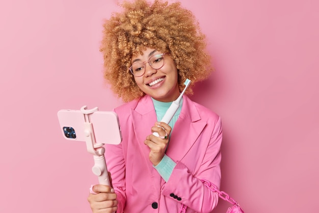 Positive, schöne, lockige Frau posiert für Selfie mit elektrischer Zahnbürste, dreht Video-Blog, gibt ihren Followern Ratschläge, wie man sich um Zähne kümmert, empfiehlt etwas, schlägt nützliche Tipps vor