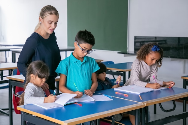 Positive Schullehrerin beobachtet Kinder, die ihre Aufgabe im Unterricht erledigen, an Schreibtischen sitzen, zeichnen und in Hefte schreiben