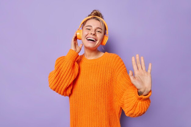 Positive sorglose Europäerin tanzt im Rhythmus der Musik, hört Lieblingslied über Kopfhörer, trägt orangefarbenen Strickpullover, lächelt breit isoliert über violettem Hintergrund, albert herum