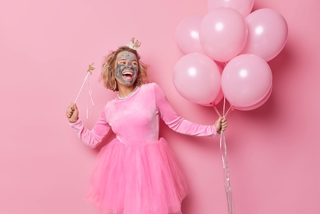 Positive sorglose Frau trägt ein festliches Kleid, wendet Tongesichtsmaske zur Hautbehandlung an, hält einen Haufen aufgeblasener Luftballons und Zauberstab singt ein Lied, das isoliert über rosafarbenem Hintergrund ist, hat fröhliche Stimmung