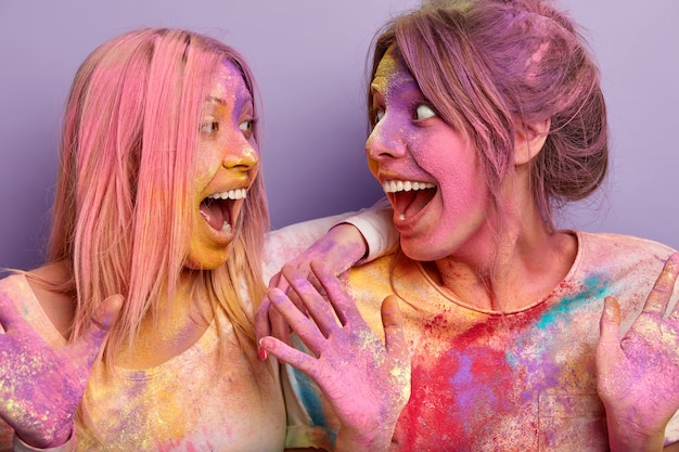 Positive überglückliche europäische Frauen sehen sich glücklich an, rufen aus und haben Spaß zusammen, schmutzig mit Holi-Farben, stehen über lila Wand, sind sehr emotional und zufrieden. Frühlingsferien