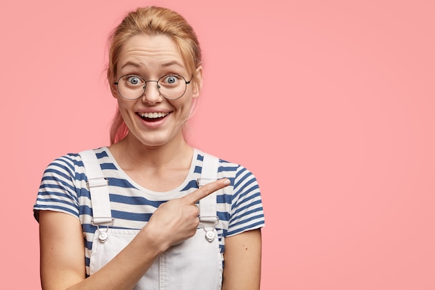 Kostenloses Foto positive verkäuferin zeigt kunden richtung, zeigt mit dem zeigefinger beiseite, gekleidet in freizeitkleidung, isoliert über rosa wand