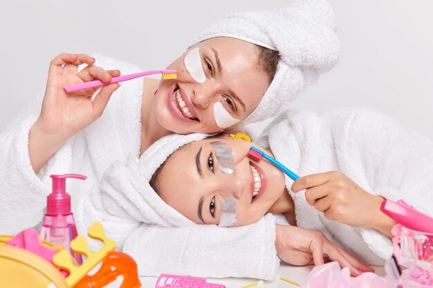 Positive vielfältige junge Frauen neigen die Köpfe angenehm lächeln kümmern sich um Teint und Zähne halten Zahnbürsten in weiche Bademäntel gekleidet Handtücher über den Köpfen werden Schönheits- und Hygieneverfahren unterzogen