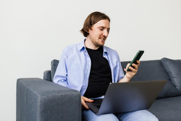 Positiver bärtiger Mann mit Laptop, der auf dem Sofa im Wohnzimmer sitzt und auf dem Smartphone spricht