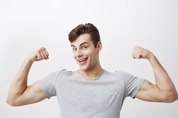 Positiver emotionaler Mann lächelt, zeigt Muskeln auf seinen Armen, ist stolz darauf, stark zu sein und Kraft zu haben, sagt: Ich bin Held. Muskulöser männlicher Athlet hebt vor Freude die Arme und zeigt, wie stark er ist.