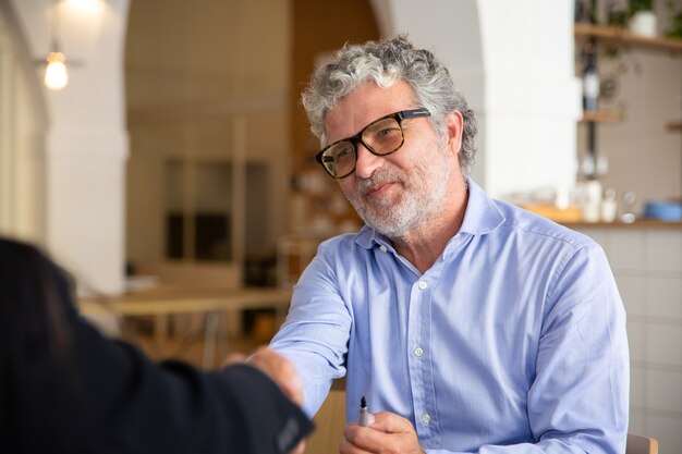 Positiver freundlicher Geschäftsmann in Gläsern, die Hand mit Partner beim Treffen schütteln