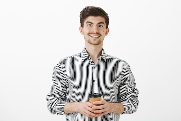 Positiver freundlicher reifer Mann mit Schnurrbart und Bart im gestreiften Hemd, Tasse Tee oder Kaffee haltend und freudig lächelnd, neue Leute im Büro treffend, lässig und sorglos über graue Wand sprechend