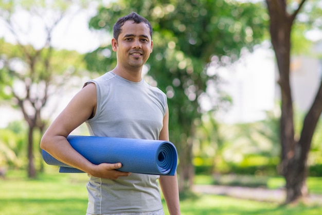 Positiver indischer Kerl mit der Yogamatte, die zur Klasse im Freien fertig wird.